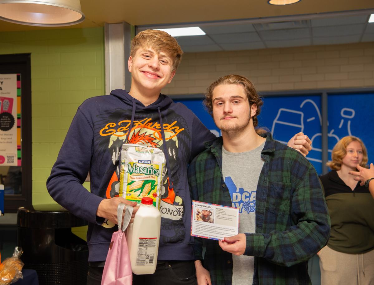 Two students smile at the camera. One is holding ingredients, and the other is holding up a recipe card.