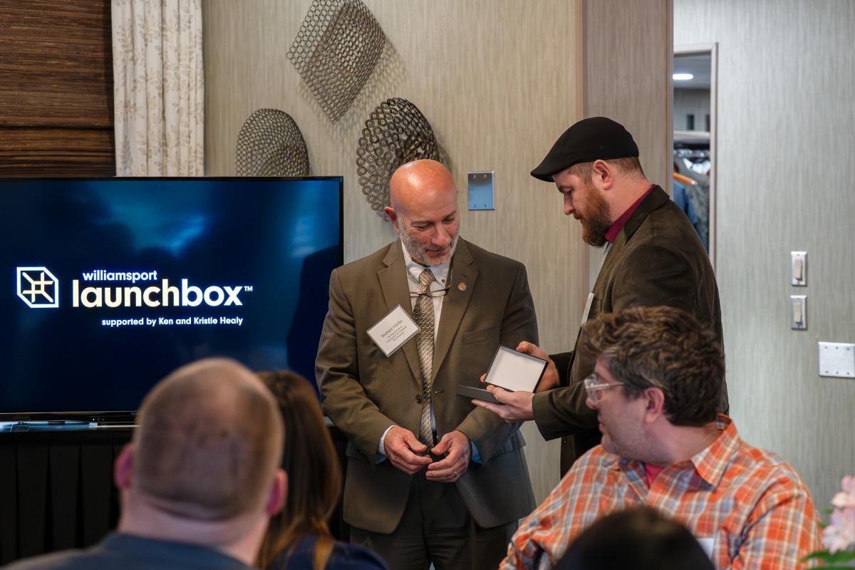 A man in business attire looks as another man opens a gift box.