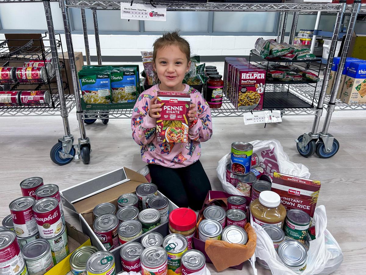 A child sits in front of shelves surrounded by canned goods and other foods and holding a box of pasta.