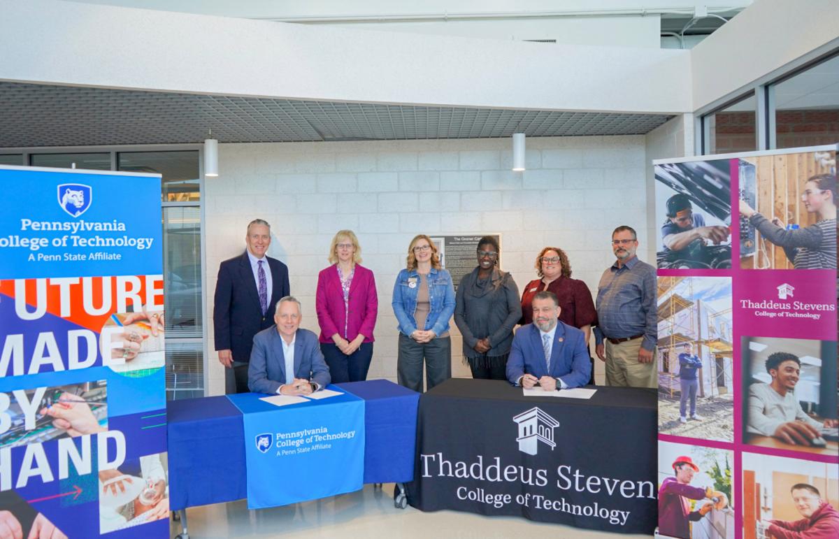 Penn College President Michael J. Reed sits behind a table with Penn College signage, and Thaddeus Stevens College of Technology President Pedro A. Rivera II sits behind a table with Thaddeus Stevens signage. A group of people from both colleges stands behind them.