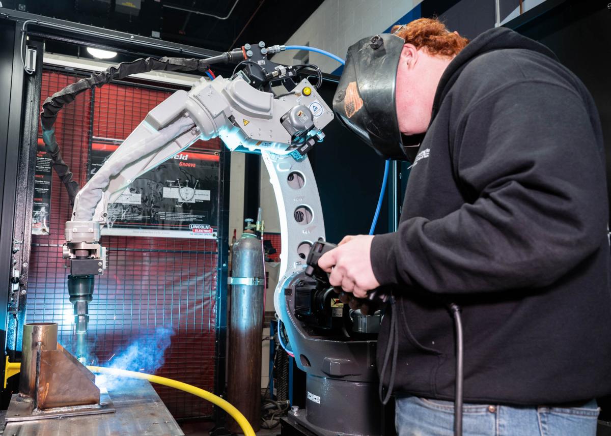 A Pennsylvania College of Technology student operates a robotic welding system. The student is wearing a welding helmet. Light and smoke emanate from the end of the robotic arm, which is touching metal.