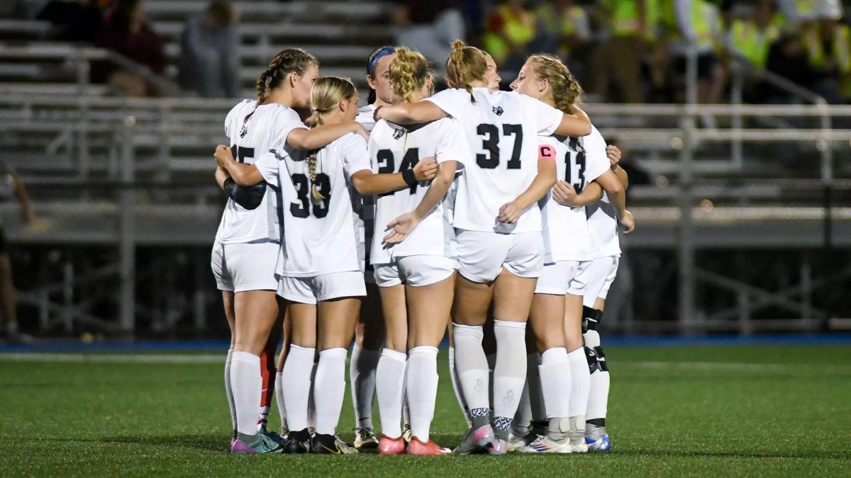 Penn College women's soccer players huddle together arm-in-arm on the field.