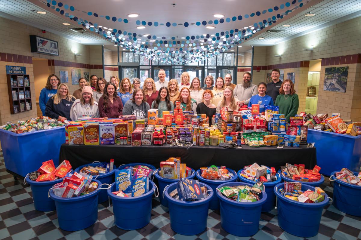 A large group of people stands behind a table filled with nonperishable food items. In front of the table are are 18 round bins filled with food, and on either side are larger bins filled with food.