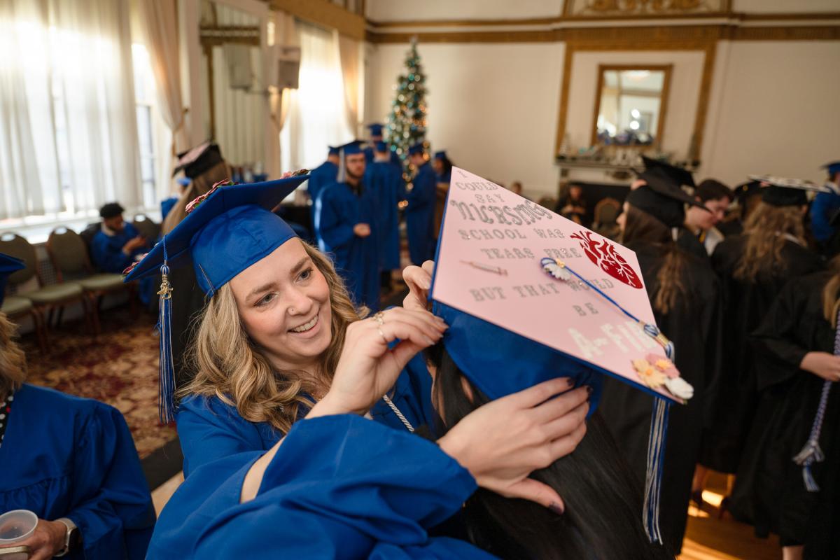 Graduate in blue cap and gown adjusting another graduate’s decorated cap, which has text and designs, in a room with other graduates and a Christmas tree in the background.