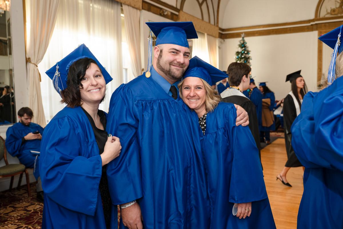Three people wearing blue caps and gowns standing together in a Genetti Hotel ballroom, with other graduates and a decorated Christmas tree visible in the background.