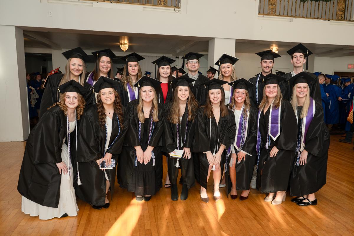 Group of graduates wearing black caps and gowns with purple stoles and honor cords posing together in a Genetti Hotel ballroom.