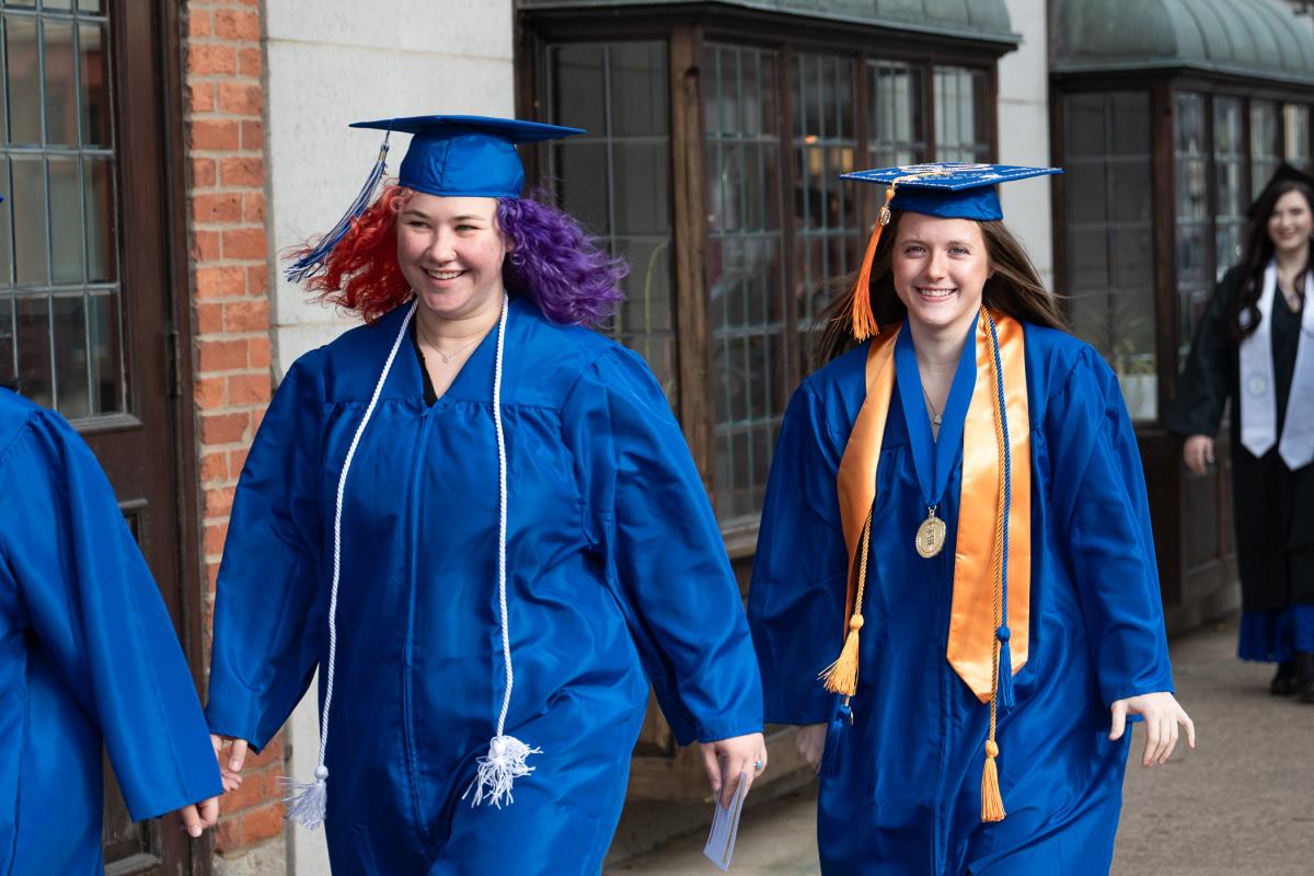 Two graduates wearing blue caps and gowns walking outside near a brick building, one with a white honor cord and the other with an orange stole and medal.