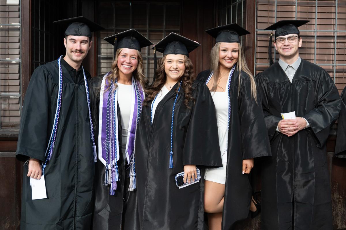 Five graduates in black caps and gowns posing together outdoors, some wearing honor cords and stoles.