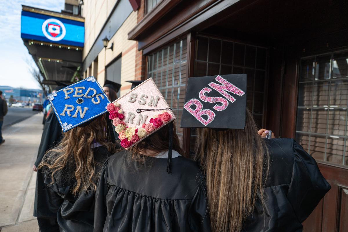 Three graduates wearing black gowns standing outside, showing decorated graduation caps with text: 'PEDS RN,' 'BSN RN,' and 'BSN.'
