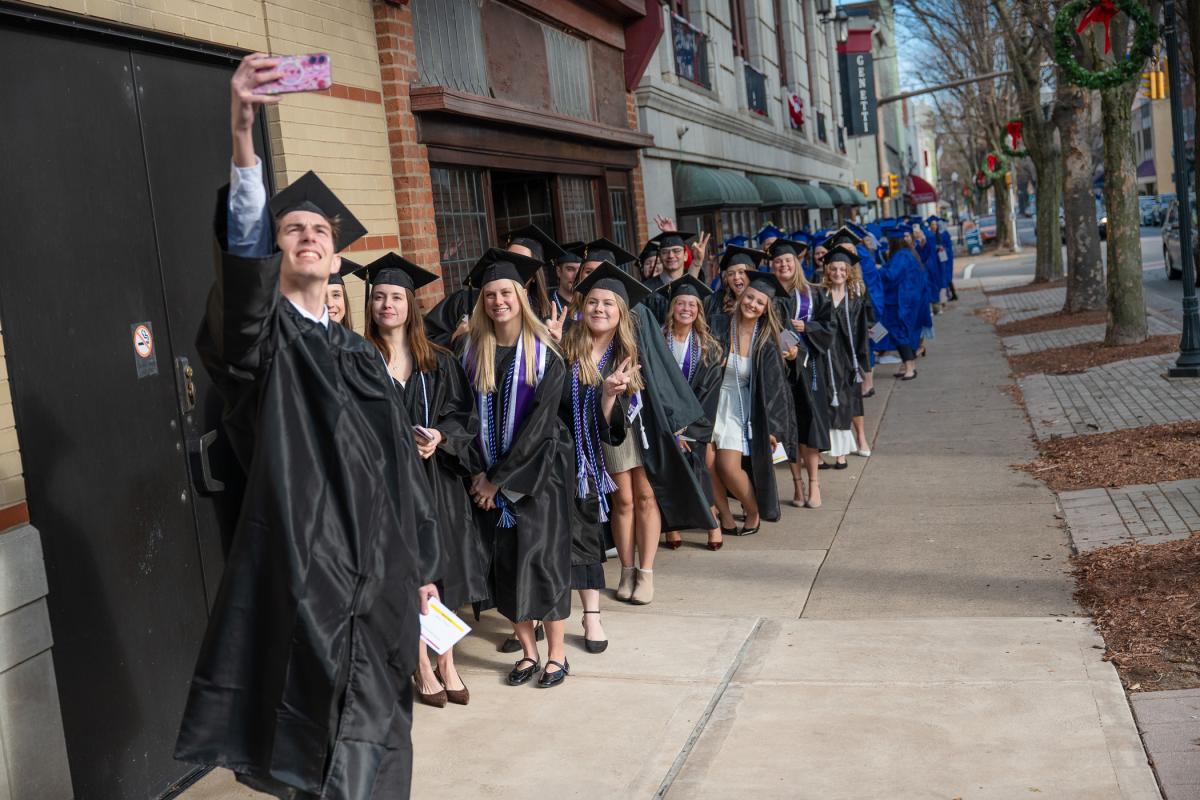 Graduates in black and blue caps and gowns standing in a line on a city sidewalk, with one person holding a phone for a selfie. Buildings and holiday wreaths visible along the street.