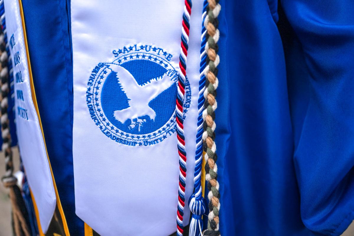 Close-up of a white graduation stole with a blue eagle emblem and the words 'Service, Academics, Leadership, Unity, Tribute, Excellence,' alongside multiple braided honor cords in red, white, blue, and tan.