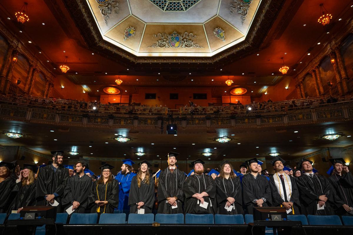 Graduates in black and blue caps and gowns standing in rows inside an ornate theater with a decorative ceiling and balconies filled with seated guests.