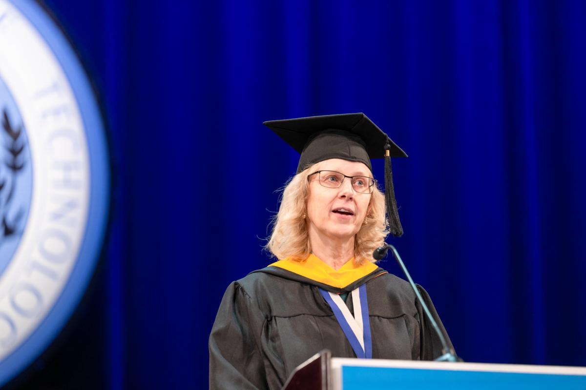 A woman wearing a black cap and gown with a gold academic hood standing at a podium on stage, with a blue curtain and a large Penn College seal in the background.
