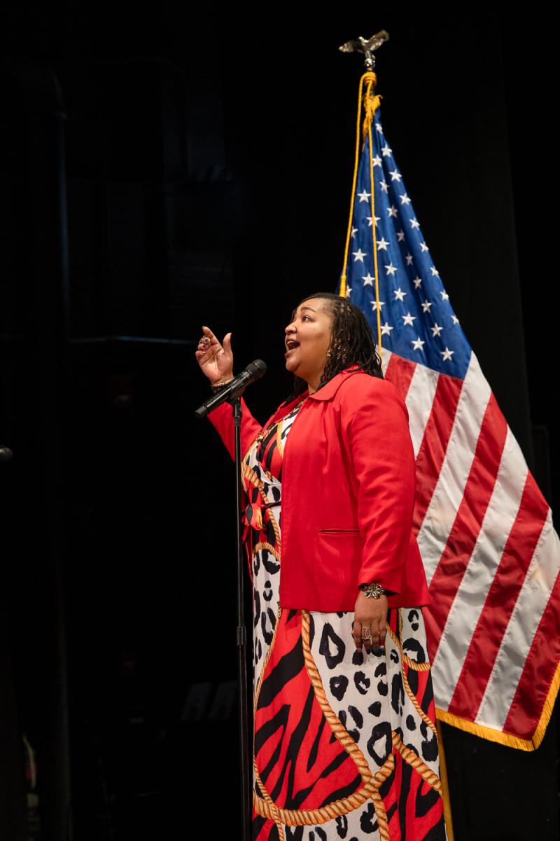 Person standing on stage wearing a red jacket and patterned dress, sings into a microphone with a large American flag displayed behind them.