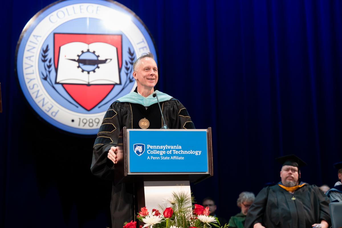Speaker at a podium with a Pennsylvania College of Technology sign, wearing academic regalia and a medallion, with a large Penn College seal and blue curtain in the background.