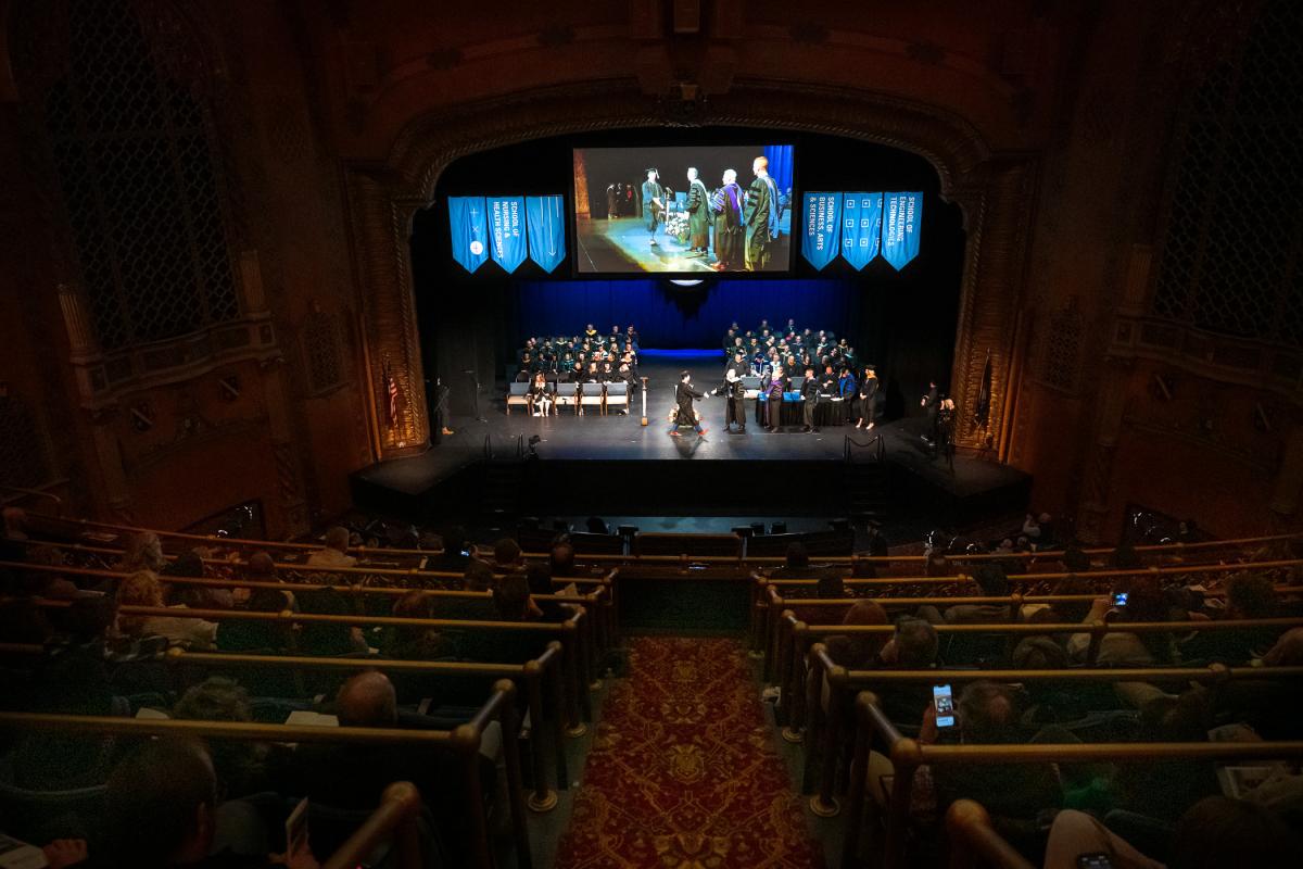 Wide view of graduation ceremony, showing graduates and faculty on stage, banners hanging above, and a large screen displaying the diploma presentation.