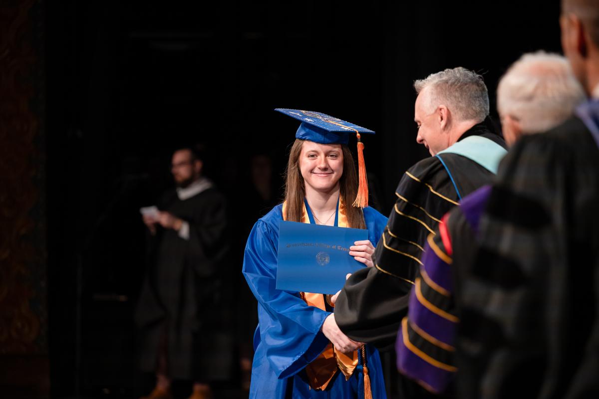 Graduate in a blue cap and gown with gold stole accepts a diploma folder from President Reed.