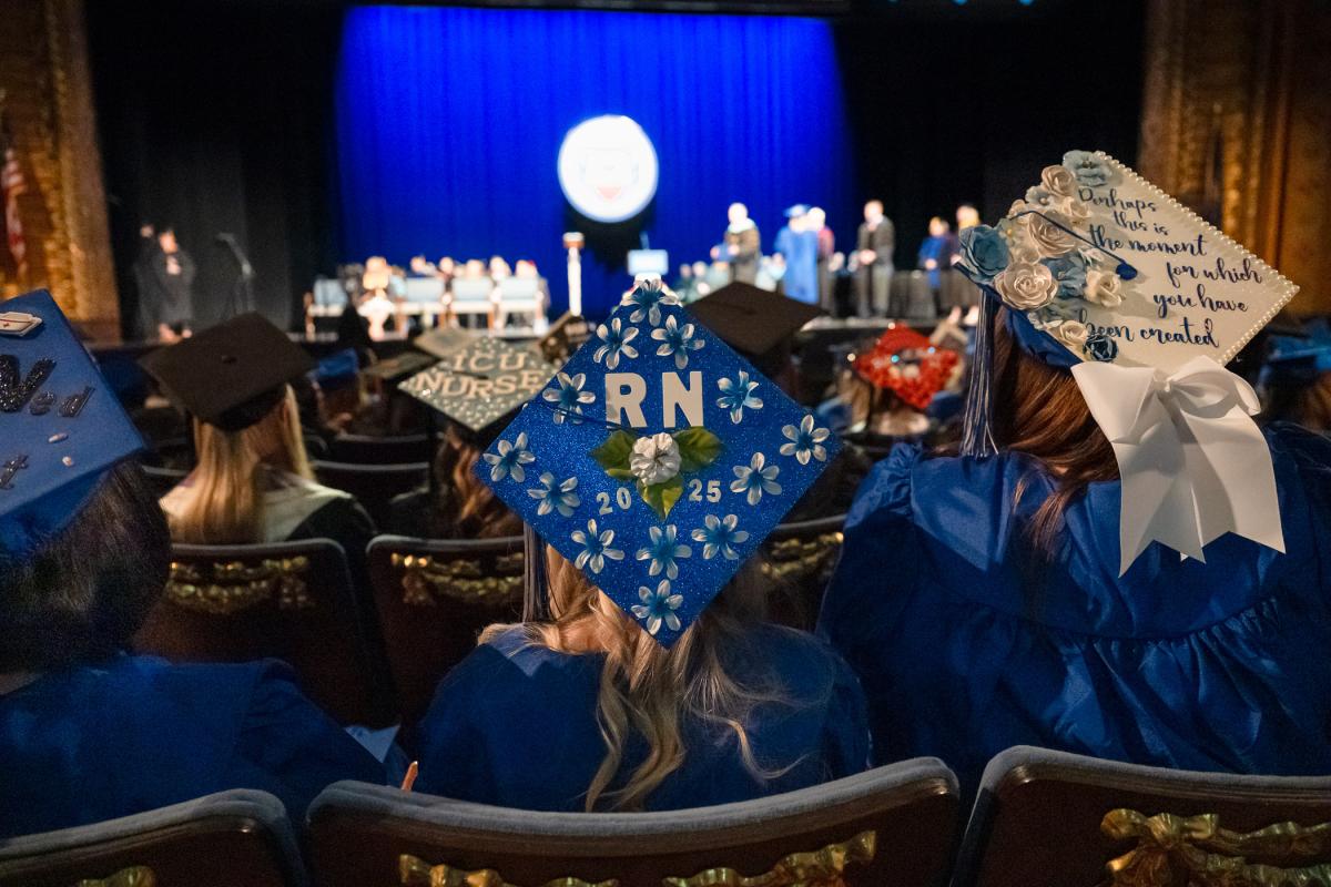 Graduates seated in the Community Arts Center wearing blue caps and gowns, with decorated caps featuring messages and designs such as 'RN 2025' and floral embellishments, while the ceremony takes place on stage in the background.