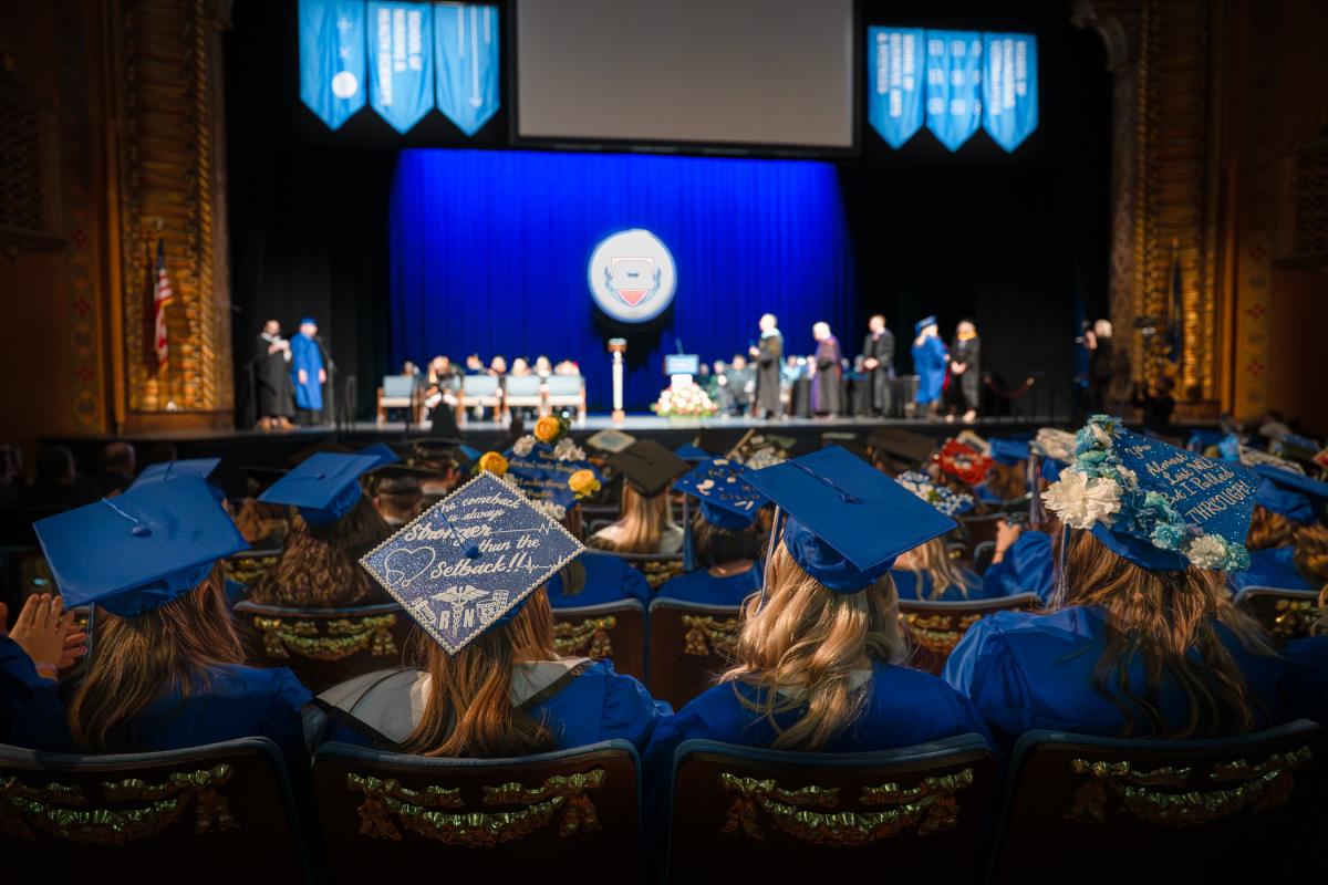 Graduates in blue gowns seated in a theater facing a stage with faculty members, decorated graduation caps visible in the foreground, and a blue curtain backdrop with banners and a podium