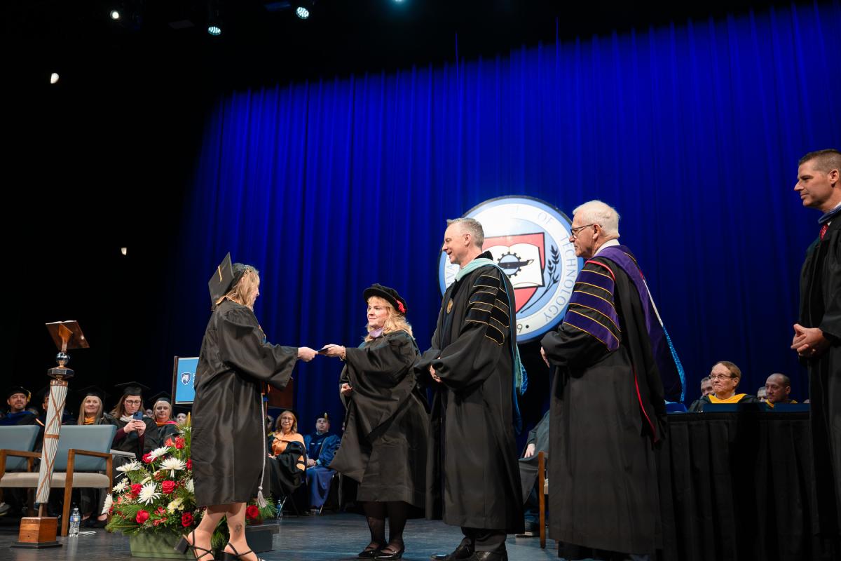 Graduate in black gown on stage receiving a diploma from President Michael J. Reed, who is wearing academic regalia, with a large Penn College seal and blue curtain backdrop behind them.