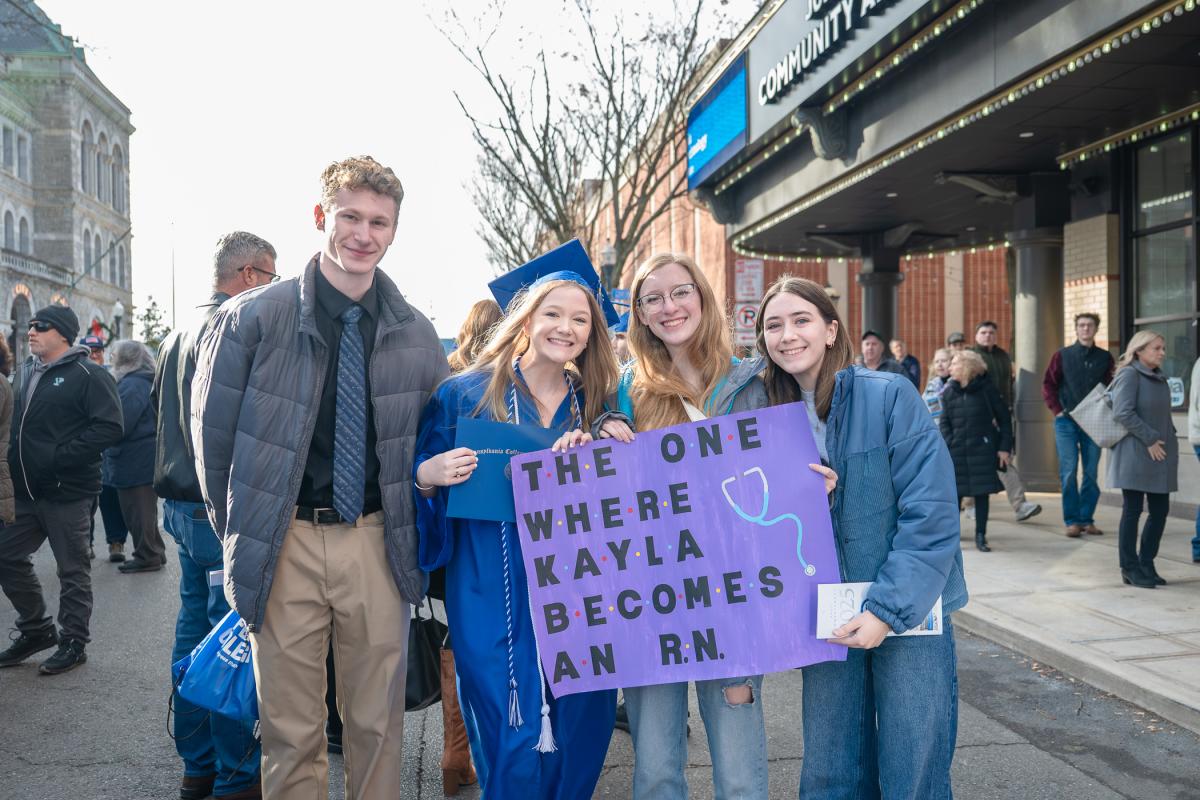 Graduate in a blue cap and gown holding a diploma folder and standing with three people, one holding a purple sign that reads 'The one where Kayla becomes an RN,' outside the Community Arts Center.