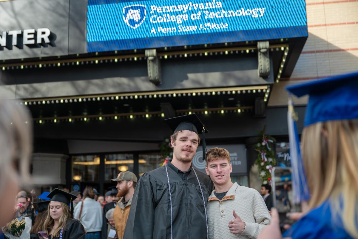 Graduate in a black cap and gown posing with another person outside the Community Arts Center, with a digital sign displaying 'Pennsylvania College of Technology.