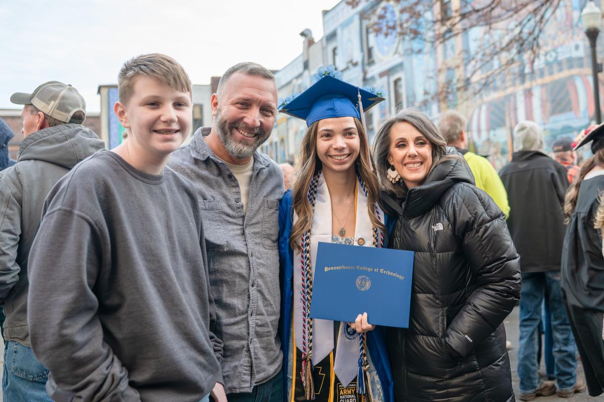 Graduate in a blue cap and gown holding a diploma folder and posing with three people outdoors in front of a mural.