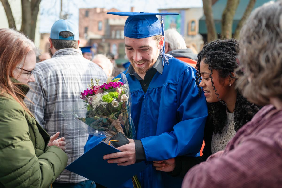 Graduate in a blue cap and gown holding a bouquet of flowers and a diploma folder while standing outdoors among a crowd of people.