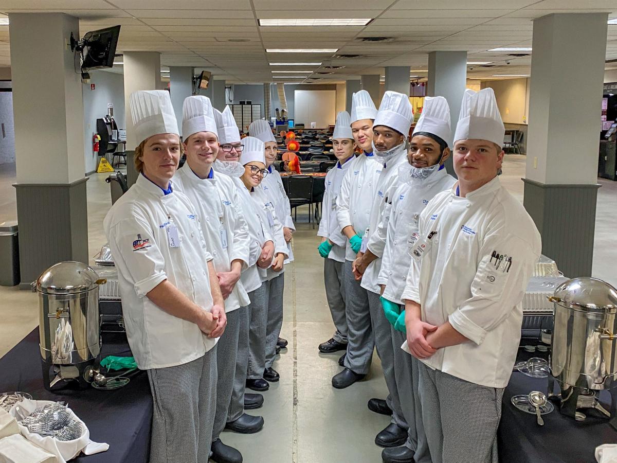 Students in chefs uniforms stand along a buffet.