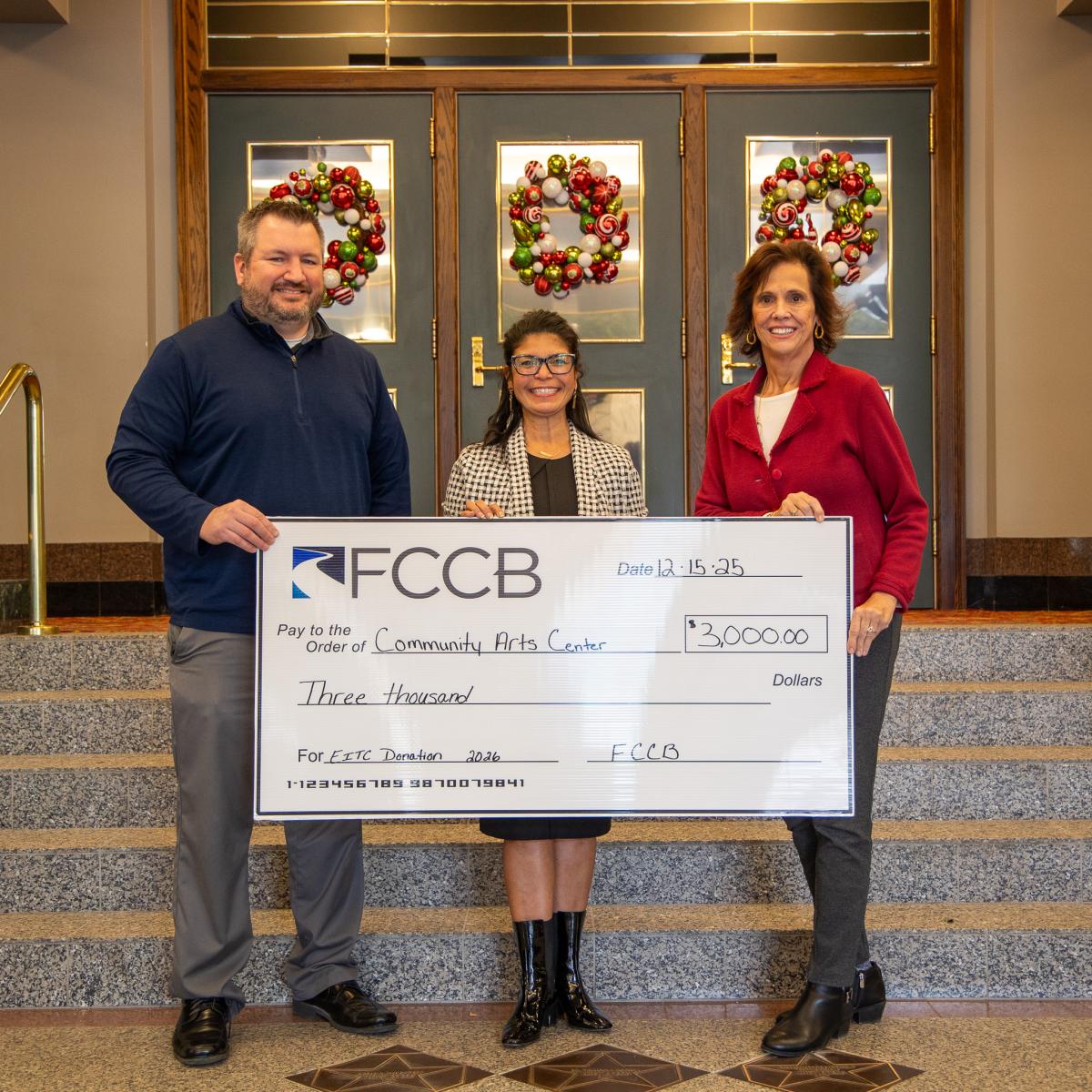Three people pose with an oversized check in the Community Arts Center lobby.