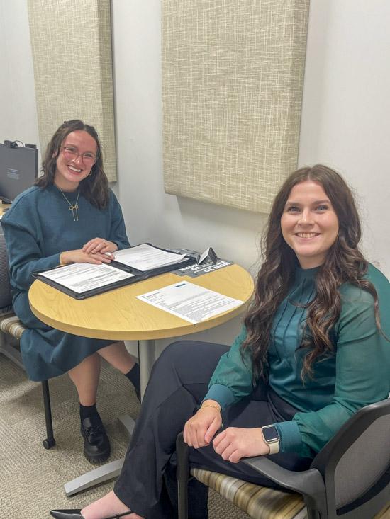 Two young women sit at a table with papers in front of them.