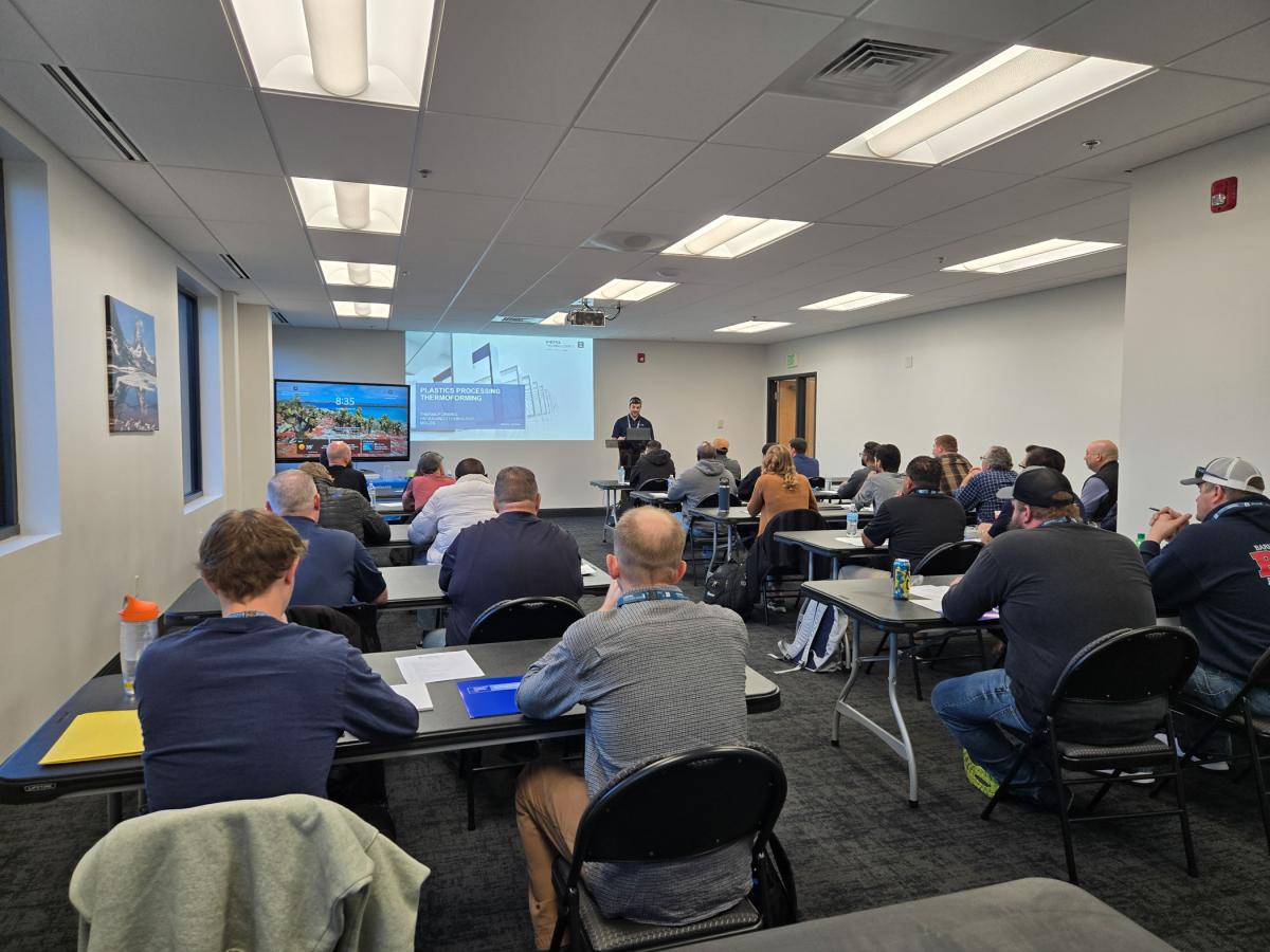Classroom setting with multiple people seated at tables facing a presenter at the front, where a large screen displays a presentation titled “Kiefel Packaging Solutions.”