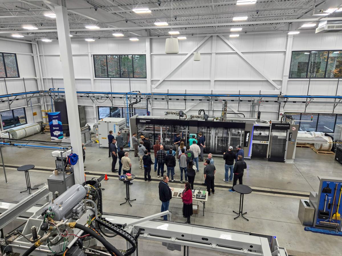 Group of people gathered inside a large industrial facility with high ceilings, observing machinery and equipment arranged along the walls.