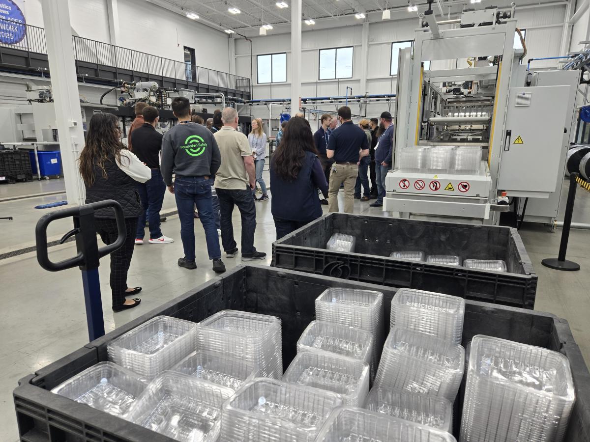  Group of people standing inside a large industrial facility near a machine producing clear plastic containers, with multiple bins filled with stacked containers in the foreground.