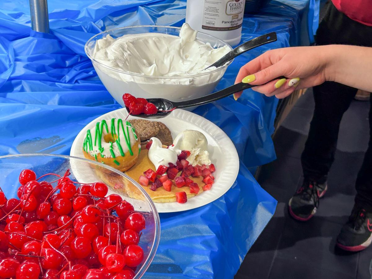 Close-up of a plate with donuts, whipped cream, strawberries, and ice cream, while a hand adds cherries from a spoon; bowls of cherries and whipped cream are visible on a blue-covered table.