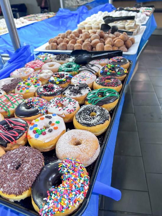 Tray of assorted decorated donuts with colorful toppings such as sprinkles, chocolate, and candy pieces, displayed on a blue-covered table.