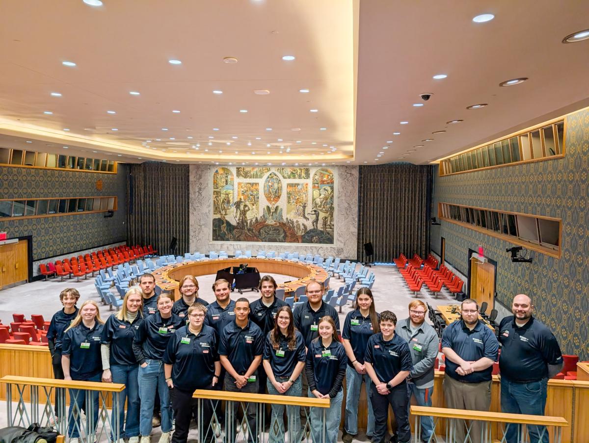 Penn College emergency management & homeland security students stand inside the United Nations Security Council chambers, which has circular seating, colorful chairs, and a large mural on the front wall.