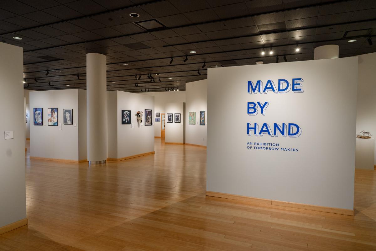 Artwork hands on walls in The Gallery at Penn College. One wall shows exhibit name, "Made by Hand," in large blue letters.