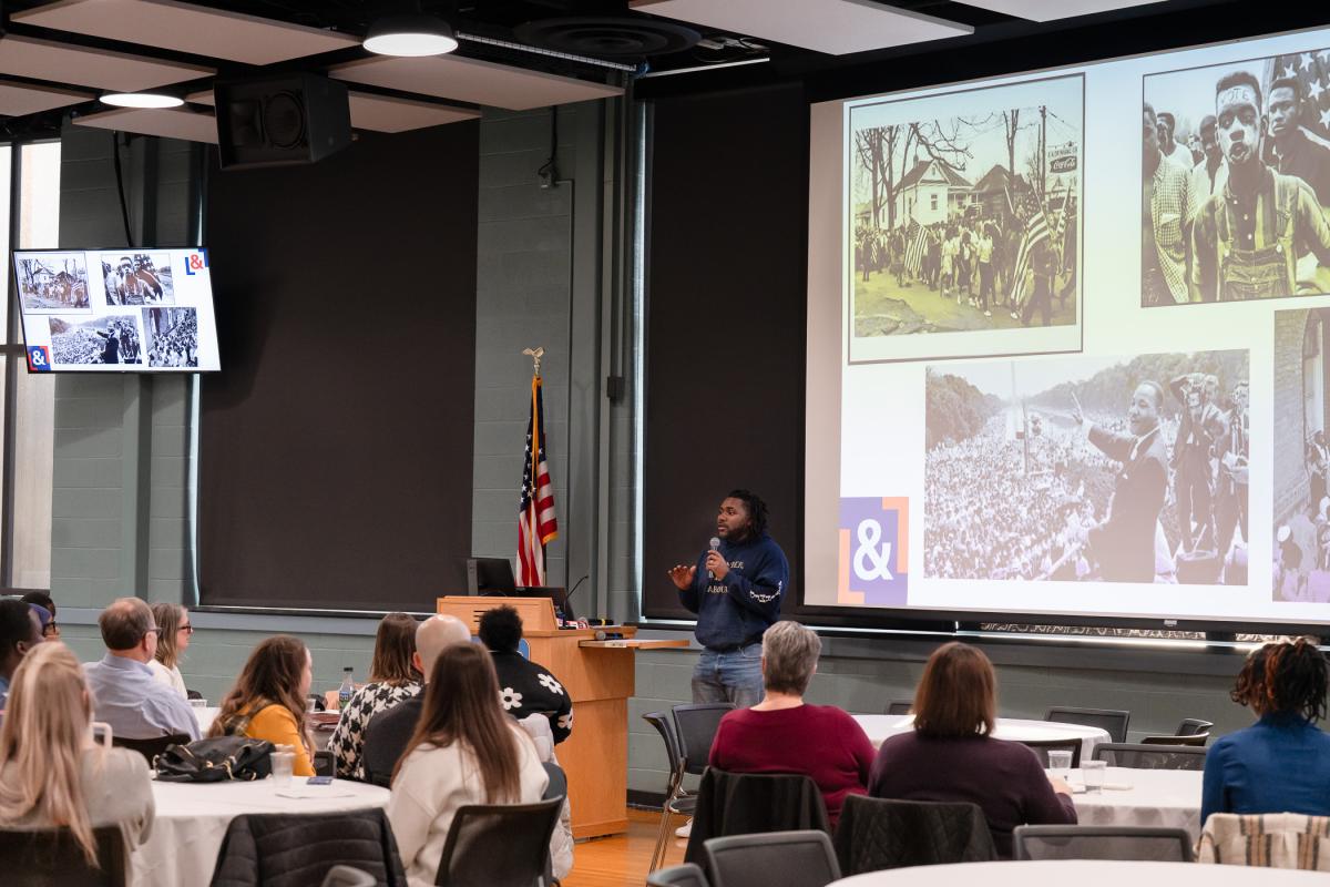 People sitting around round tables in Penn College's Penn's Inn look at a large projection screen that shows photos of Martin Luther King Jr. and the Civil Rights movement, and a man stands near a podium with a microphone.