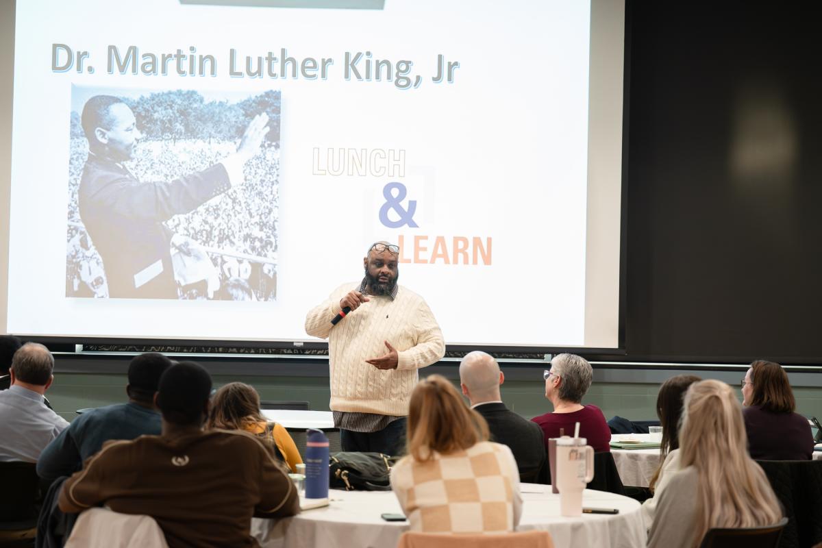 A man with a microphone stands in front of a group of people seated at round tables.