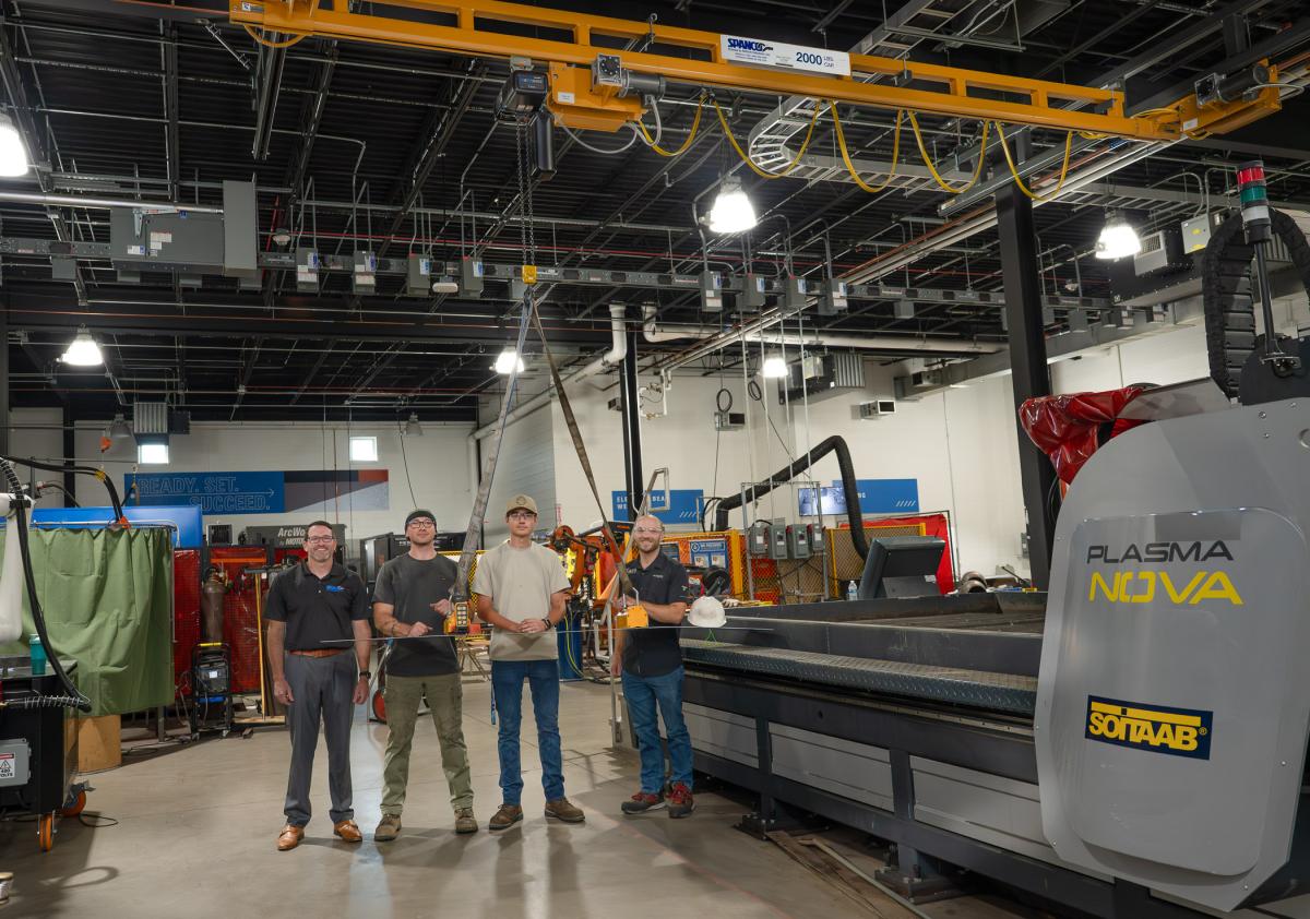 Four people standing in Penn College’s Lycoming Engines Metal Trades Center beneath an overhead crane, surrounded by welding equipment, fabrication tools, and a plasma cutting machine.