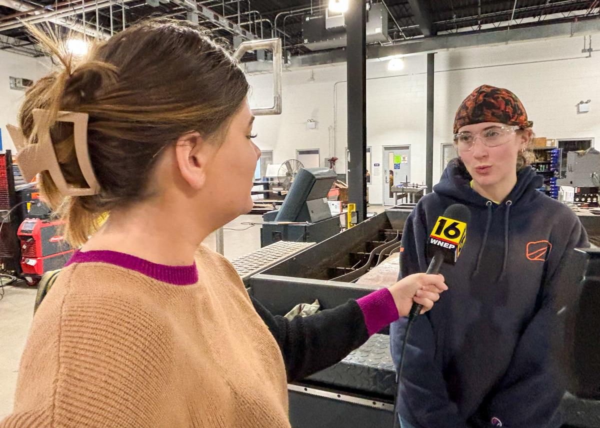A woman holds a microphone to another woman. They are surrounded by manufacturing equipment in Penn College's Lycoming Engines Metal Trades Center.