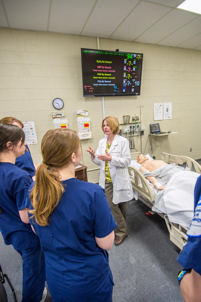 Penn College nursing students in blue scrubs look at an instructor who is wearing a white lab jacket and standing near a medical manikin in a hospital bed.