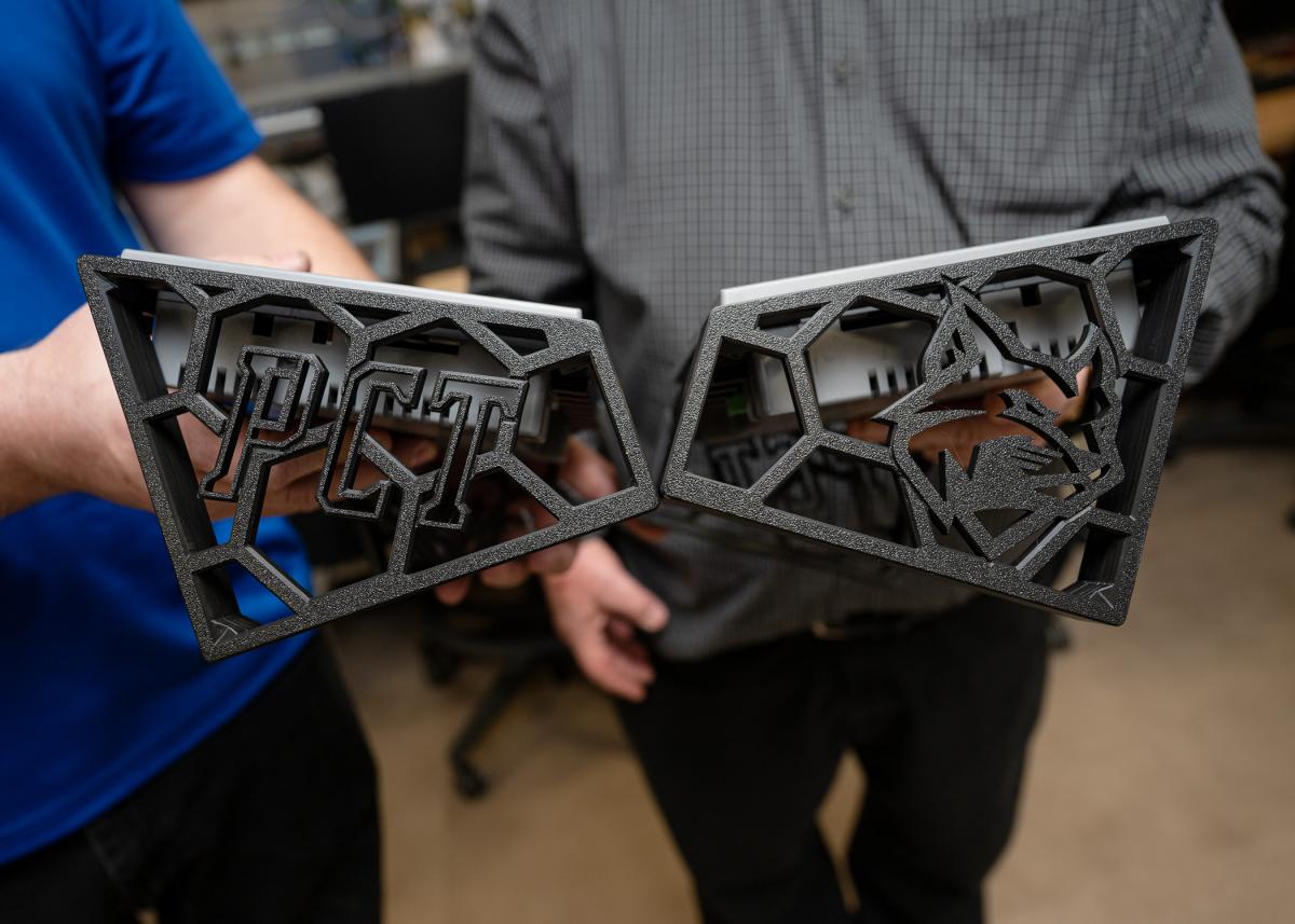 Close-up of hands holding right-trapezoid-shaped human machine interface frames. One shows the left side, with large "PCT" letters, and the other shows the right side of the frame, with the college's Wildcat logo. 