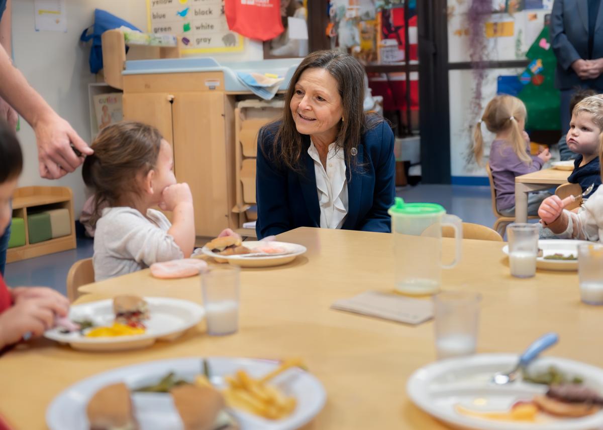 A woman sits at a table and chats with a child who has a plate of food in front of her.