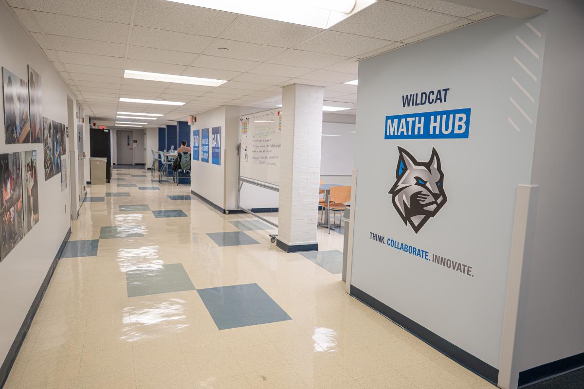 A wall at the entry of an open space with tile floors, tables and chairs and white boards has a Wildcat logo and says "Wildcat Math Hub. Think. Collaborate. Innovate."