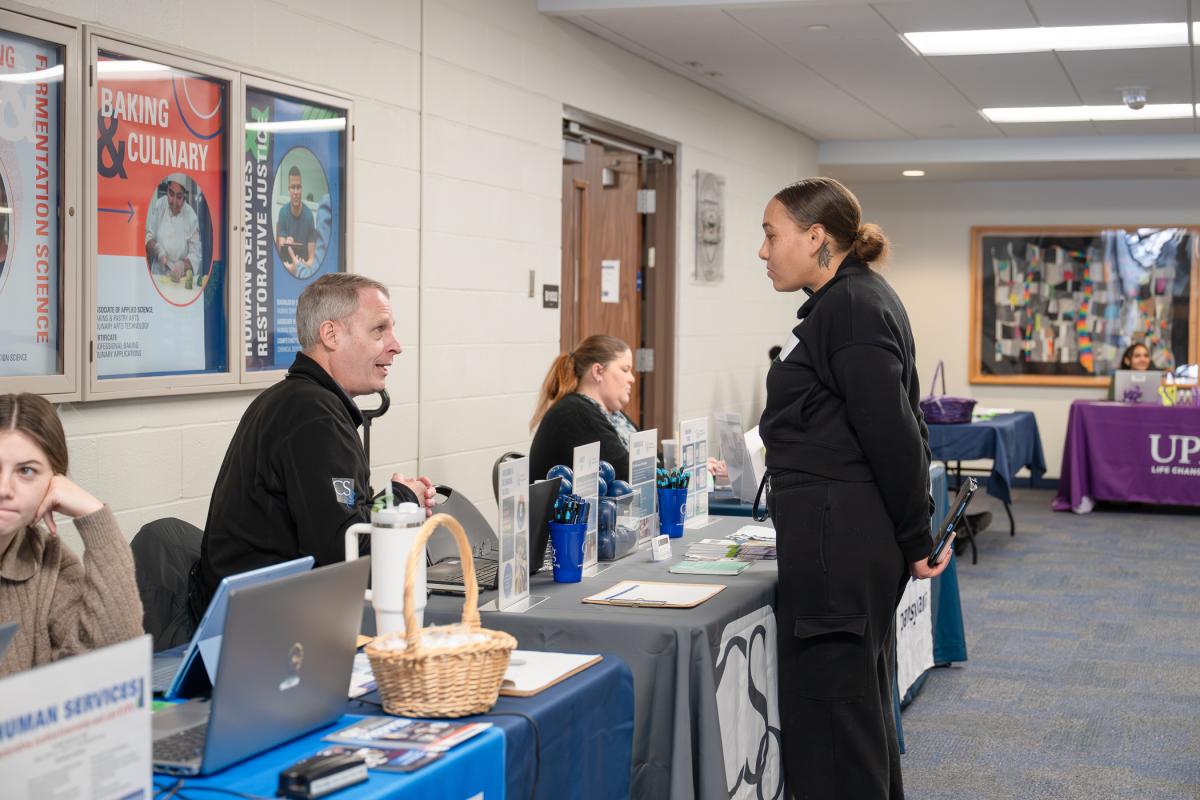 A man sitting behind a table covered with logos, pamphlets and business cards talks with a student standing on the other side.