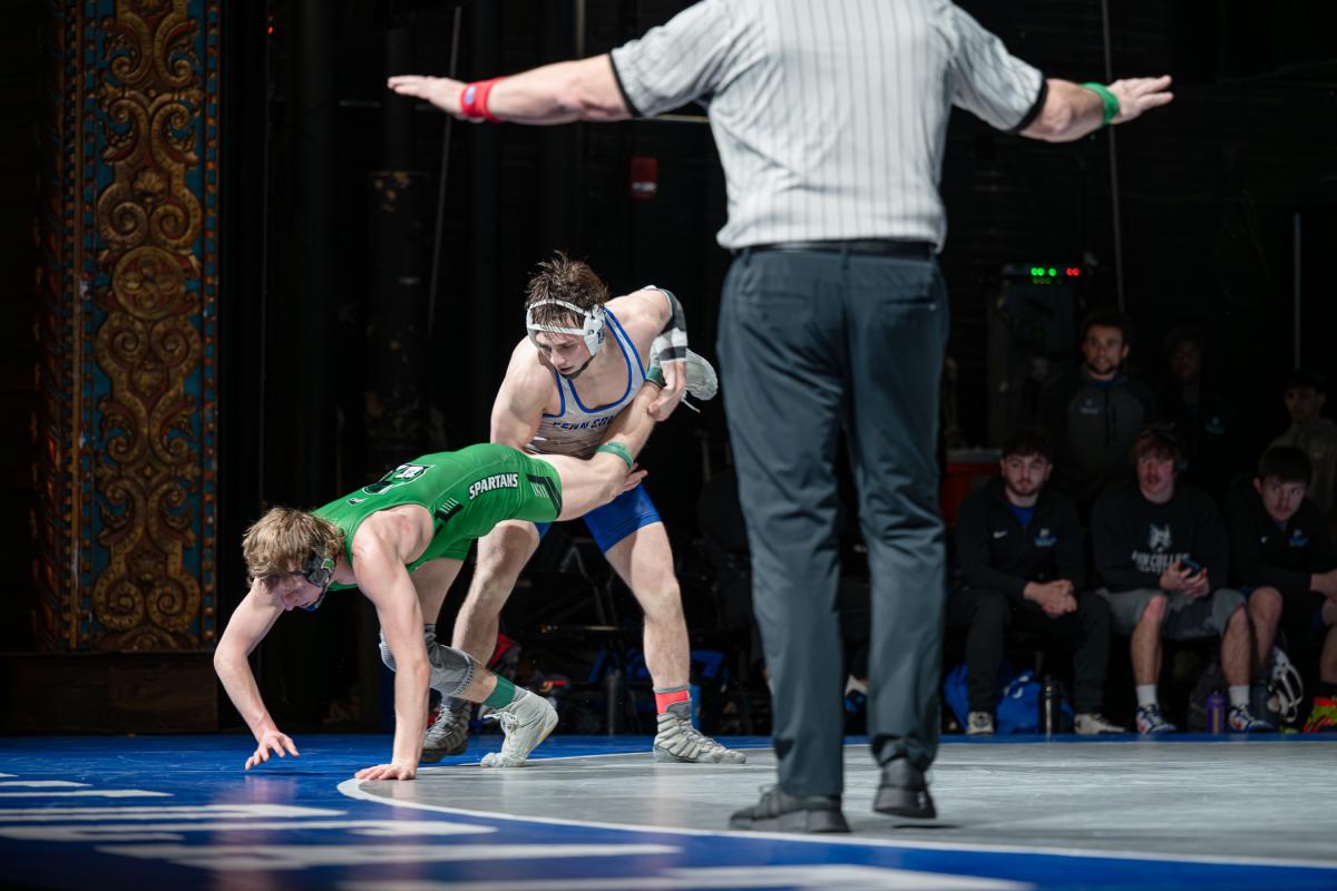 A wrestler in green attempts to escape while an opposing wrestler applies a standing hold, as a referee signals nearby during a match.
