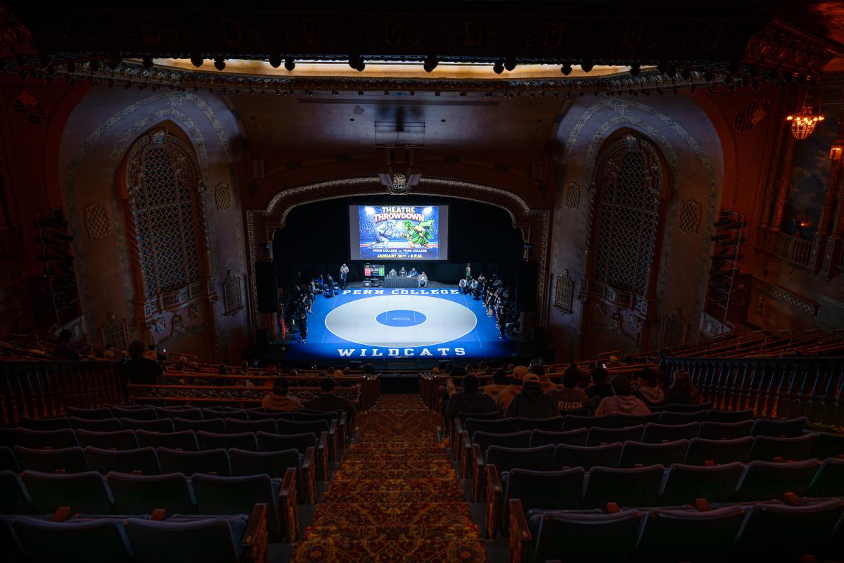 Audience perspective of a wrestling match in progress, with the mat centered on the stage of an ornate theater and spectators seated throughout the theater.