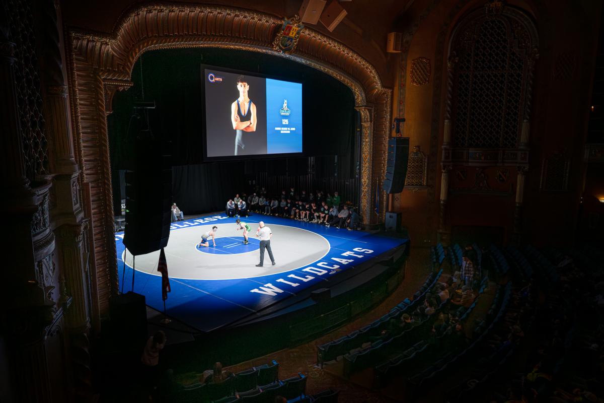 High, wide-angle view of a wrestling match taking place on a stage inside an ornate theater, with seating visible around the mat.